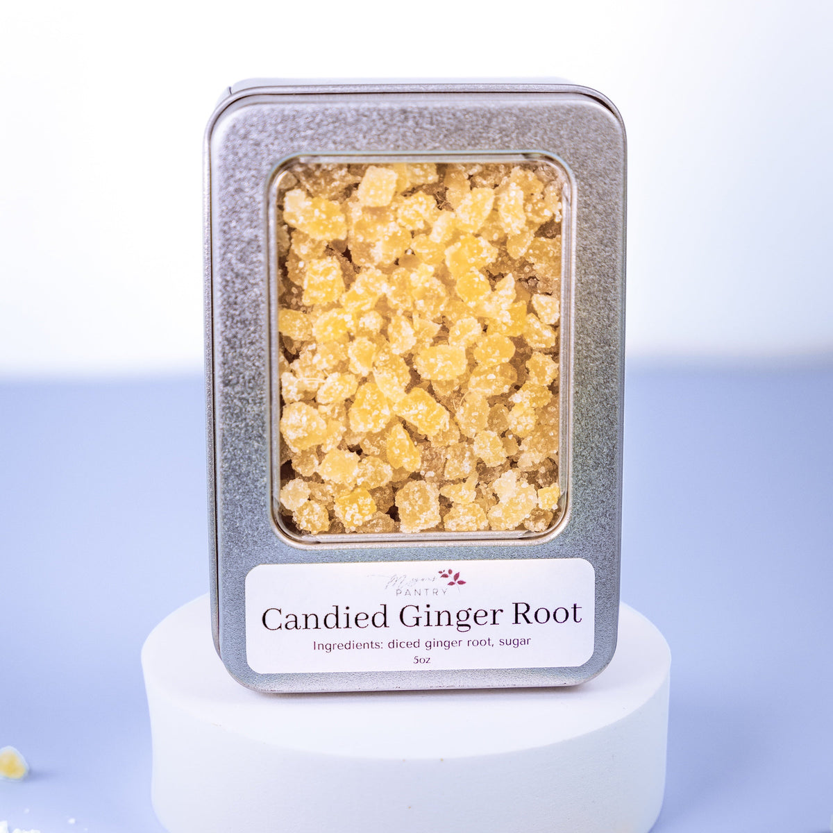 Candied ginger root in a metal tin on a white pedestal with a light blue background