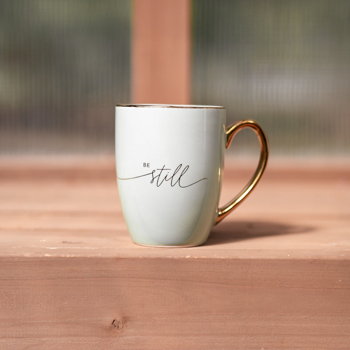 Sage green ceramic mug from Megan's Pantry with a gold rim, gold handle, and "be still" lettered in gold.